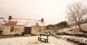 Image shows snow covered ground, a bench seat and a snow covered red roofed stone cottage (painted white a long time ago)